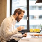 a man working on a laptop sitting on a chair having a cup of coffee in his left hand referencing it as a picture for resolving and identifiying Nacha file errors